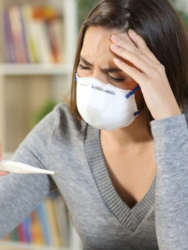 IMAGE: A sick woman wearing a mask looking at a thermometer